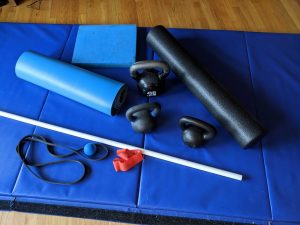 Image of Ren's at-home workout equipment. Shows three kettlebells, long foam roller, foam stability pad, yoga mat, one long band and one circle band, a lacrosse ball, and a PVC pipe on top of a thick blue gymnastics pad.